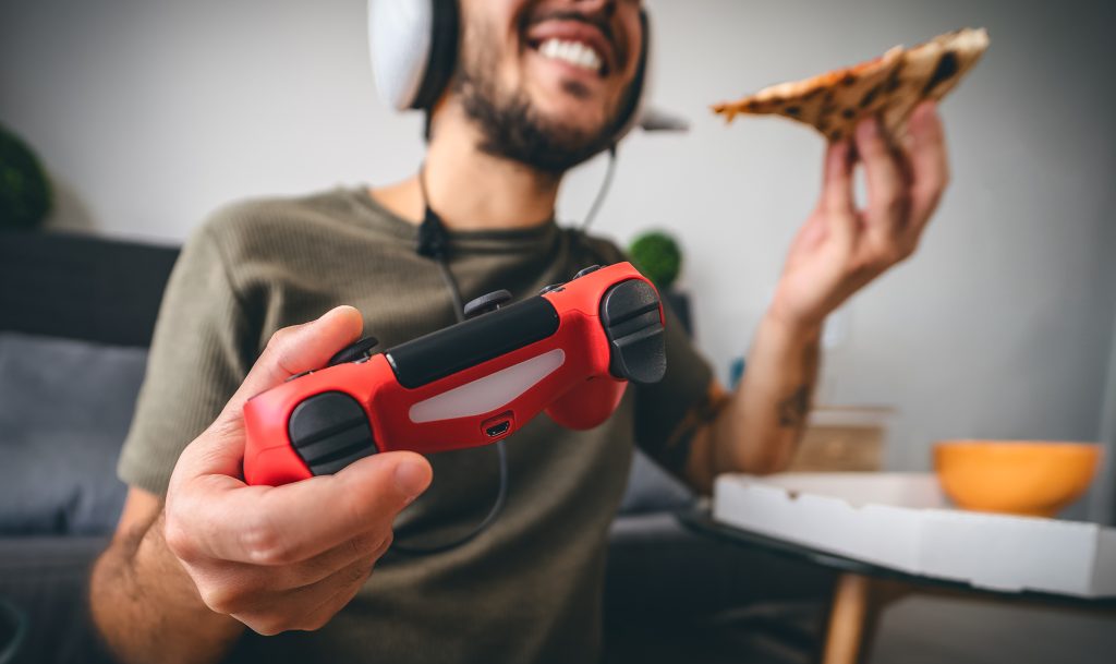 Homem jogando video-game com controle vermelho e headset segurando um pedaço de pizza