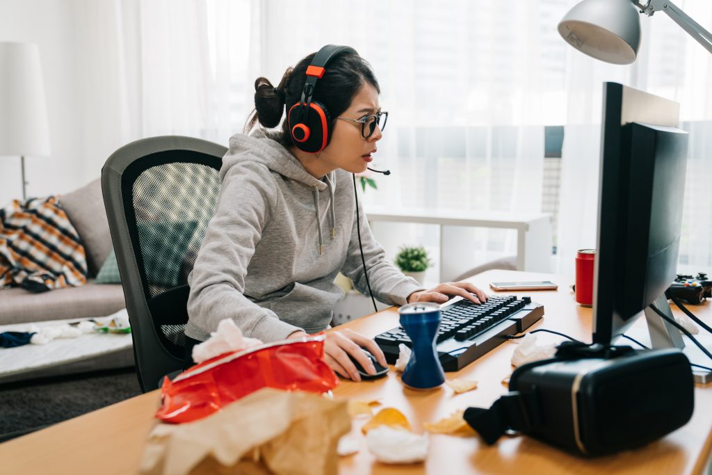 Mulher jogando video-game no computador com headset em mesa com uma latinha amassada e um saco de snack representando o hábito de consumo dos gamers quanto à comida e bebida