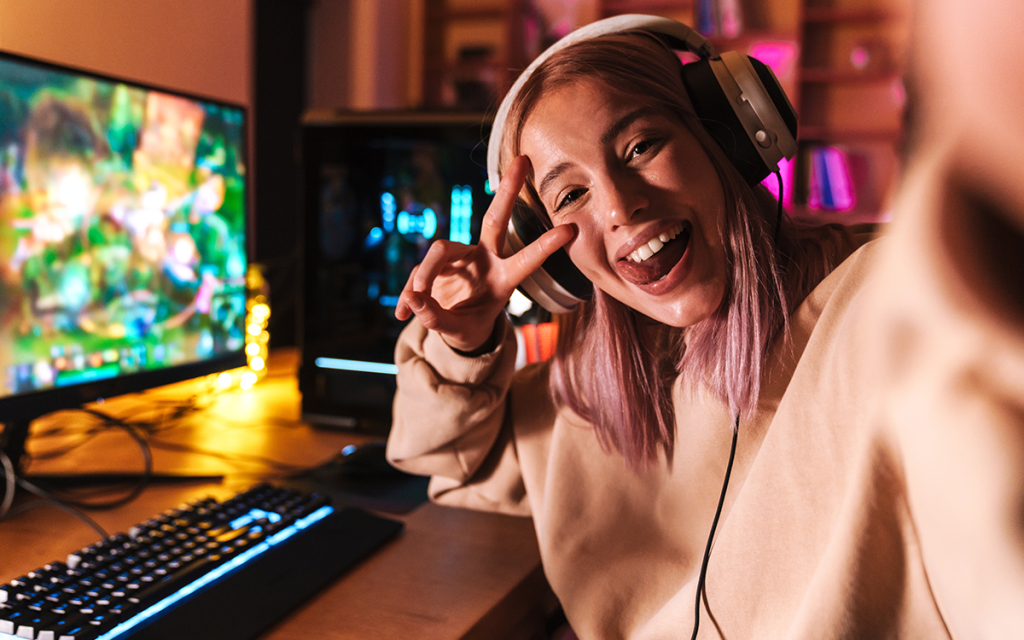 Garota gamer tirando selfie fazendo a letra V com a mão ao lado de computador com teclado e headphone