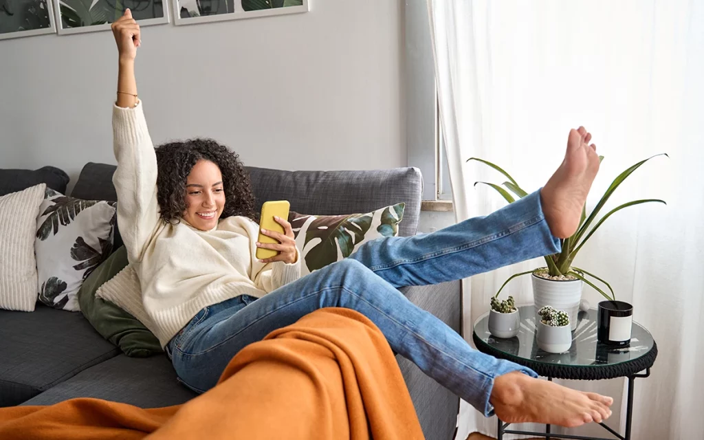 Uma mulher alegre comemorando uma vitória enquanto olha para seu smartphone em casa.