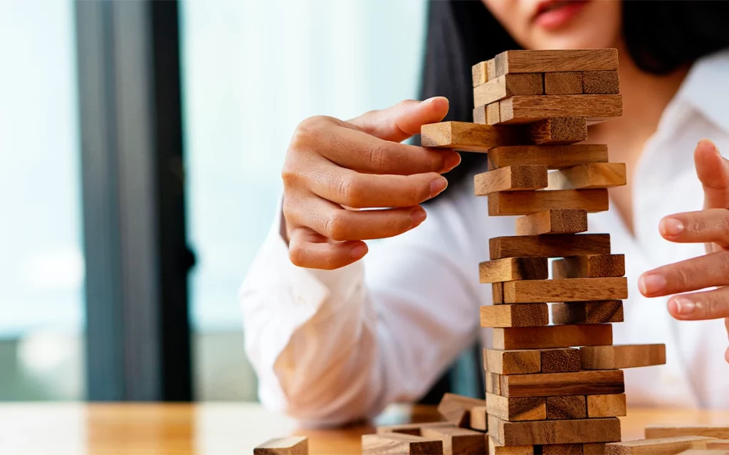 Mão feminina cuidadosamente movendo um bloco em uma torre de jogo Jenga, destacando o equilíbrio e a estratégia, elementos comuns em jogos utilizados na gamificação.