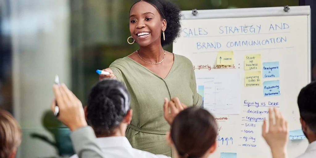 Mulher entusiasmada conduzindo uma sessão de treinamento e desenvolvimento com um quadro branco ao fundo, detalhando estratégias de vendas e comunicação de marca.