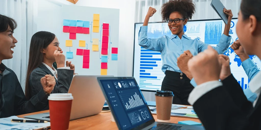 Equipe em uma sala de reuniões animada durante treinamento e desenvolvimento, com uma mulher de cabelos cacheados em destaque, demonstrando sucesso diante de gráficos e dados no quadro.